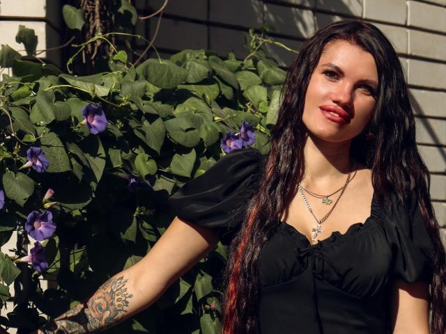 MilaLoona with long dark hair, red lipstick, black dress, puffy sleeves, tattoos on right arm, necklace, standing by green plant with purple flowers, sunlight, smiling.