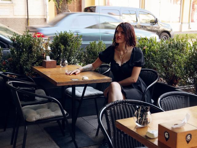 MilaLoona, with red lipstick and black dress, sits at a wooden table in a café, her hand on the table, showing her white glove, and her ass slightly visible.