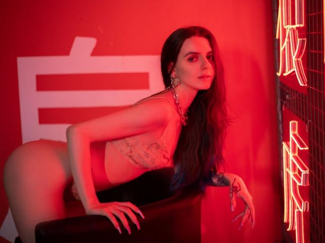 Yorogumo with long dark hair, wearing a sheer lace top and red thong, leaning on black furniture, red neon lights, and large earrings, showing her tits and ass.
