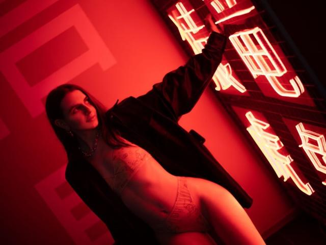 Yorogumo with long dark hair, wearing an open black jacket, sheer lace top, and high-waisted panties, standing against neon red signs, looking seductive with a slight smile.