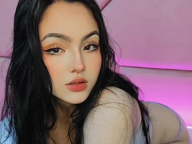 HaleiBeckham has long black hair, orange eyeliner, and pink lips, staring at the camera with a serious expression, showing her bare shoulder and a hint of her tits.