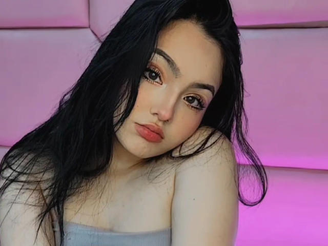 HaleiBeckham, con lunghi capelli neri, ombretto rosa e labbra rosse, appoggiata sulla spalla, indossa un top grigio senza spalline, sfondo illuminato di rosa, guarda verso la fotocamera..