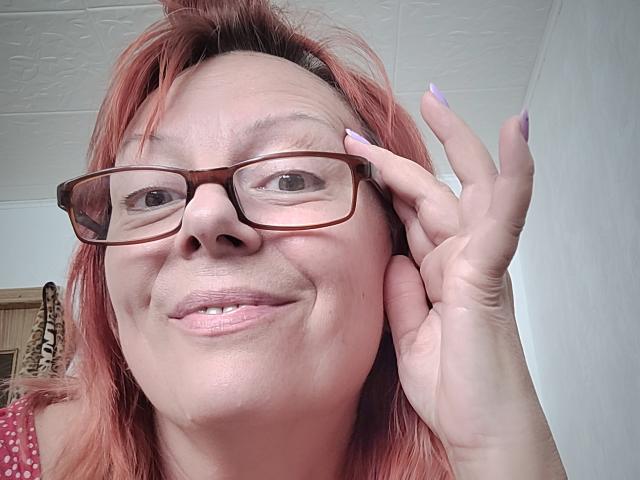 BarbaraBowie with red hair, glasses, and a smile, holding her cheek with a purple-nailed hand, showing her nose and lips. Her tits are not visible.