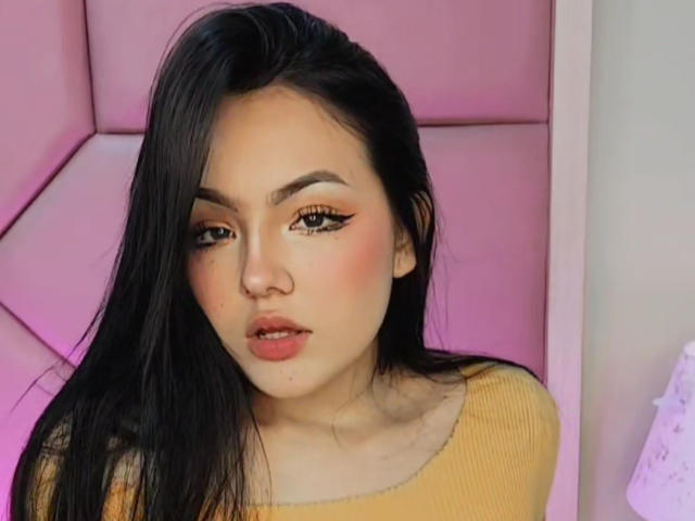 HaleiBeckham ha lunghi capelli neri, occhi marroni con eyeliner arancione, guance rosate e indossa una maglietta gialla a coste, guardando leggermente verso il basso con un'espressione dolce..