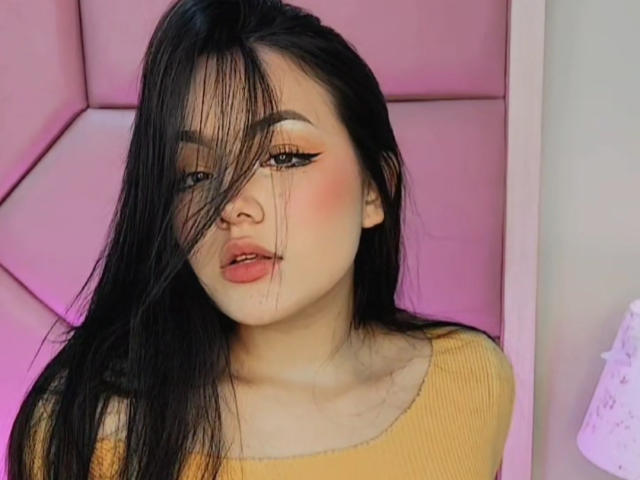 HaleiBeckham mit langem, schwarzem Haar, rosafarbenen Wangen, Eyeliner im "Flügel"-Stil und einem gelben, gerippten Oberteil, wobei ihr Haar teilweise ihr Gesicht bedeckt, und einem dezent rosafarbenen Hintergrund..