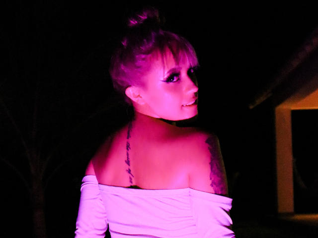 SabrinaCole, com cabelo iluminado em tons de rosa, blusa branca com um ombro descoberto, tatuagem na lâmina direita da escápula, olhando por cima do ombro, sorrindo, fundo escuro, casa visível à direita..