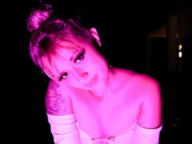 SabrinaCole, con i capelli raccolti in uno chignon e leggermente colorati di rosa, guarda verso l'alto con un sorriso appena accennato, mostrando il seno in una maglietta bianca con una spalla scoperta, rivelando un tatuaggio sulla spalla destra..