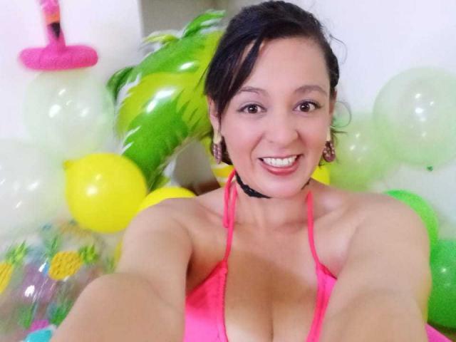 AmandaHotL with dark hair in a pink bikini top, smiling, holding a selfie stick, surrounded by balloons and a green inflatable. Her boobs are visible, she's cute.