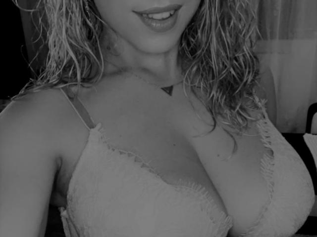 Foto em preto e branco de LucilleXSugar com cabelo loiro ondulado e molhado, usando um sutiã de renda que mostra seus seios grandes e arredondados, uma tatuagem discreta acima do colo, um sorriso leve..