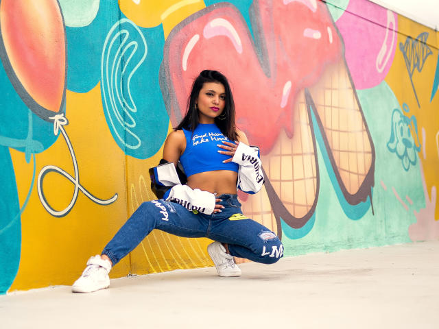 Kristal Jacobs está agachada, vestindo uma camiseta cropped azul, jeans rasgados e tênis brancos, com um casaco jogado sobre os ombros, mostrando seus abdominais definidos e seios de tamanho médio..