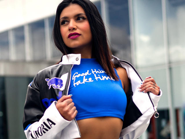Kristal Jacobs, com cabelo escuro, batom rosa, blusa cropped azul que mostra os seios, jaqueta branca e preta, com a frente aberta, exibindo o logo da Red Bull, está em pé do lado de fora..