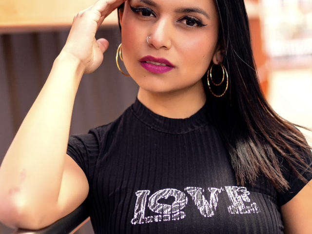 Kristal Jacobs, com cabelo preto e liso, batom roxo, brincos de argola dourados, piercing no nariz, camisa preta texturizada, mão tocando a cabeça, pele morena, atraente, contorno dos seios visível, expressão confiante..