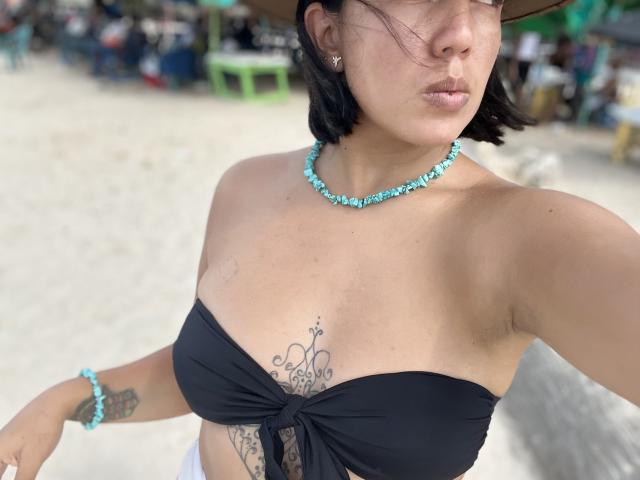 MartinaSantorin mit kurzen, schwarzen Haaren, trägt ein schwarzes Top ohne Träger, eine blaue Perlenkette und ein passendes Armband. Man sieht ein Tattoo unter ihrer Brust und sie hat leicht geschürzte Lippen. Im Hintergrund ist ein Strand zu sehen..