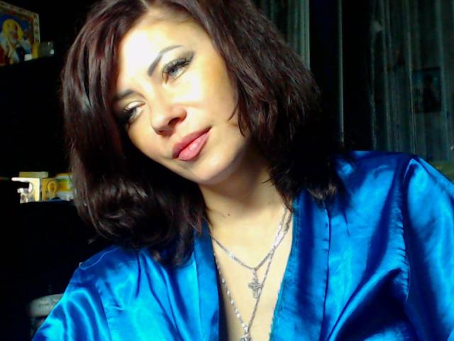 JuliaIce, avec ses cheveux bruns ondulés, portant une robe en soie bleue brillante, a les lèvres roses, un collier avec une croix et un léger sourire, et regarde sur le côté..