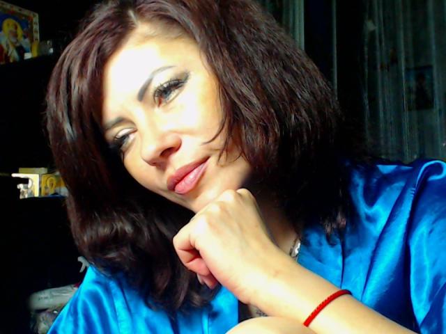 JuliaIce, com cabelo castanho escuro ondulado, camisa de cetim azul e pulseira vermelha, apoiando o queixo na mão, sorrindo suavemente com os lábios rosados ligeiramente entreabertos..