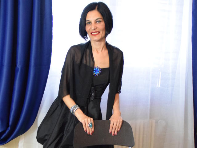 KalystaGreen, con cabello corto y negro, vestido negro, mangas transparentes, collar con una flor azul, lápiz labial rojo, sonriendo, cortinas azules, cortina blanca, silla negra, sosteniéndola con las manos en la parte superior..