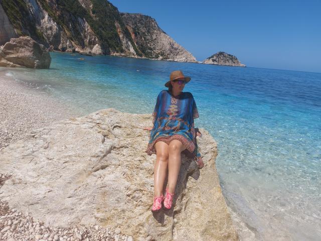 CurvySweetGirl sits on a rock, wearing a blue patterned dress, brown hat, and pink floral sandals, with her legs crossed and a calm, clear blue ocean behind her.
