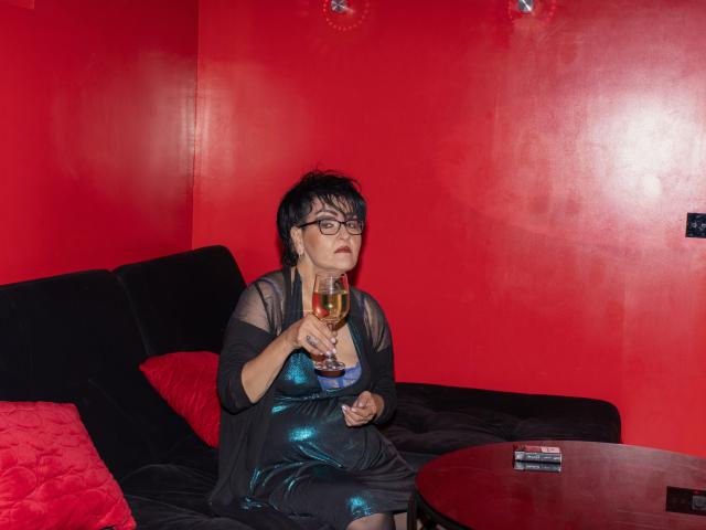 SondraRiddle with black hair, glasses, and black sheer dress, holding a glass of wine, sitting on a black couch with red pillows, in a red-walled room.