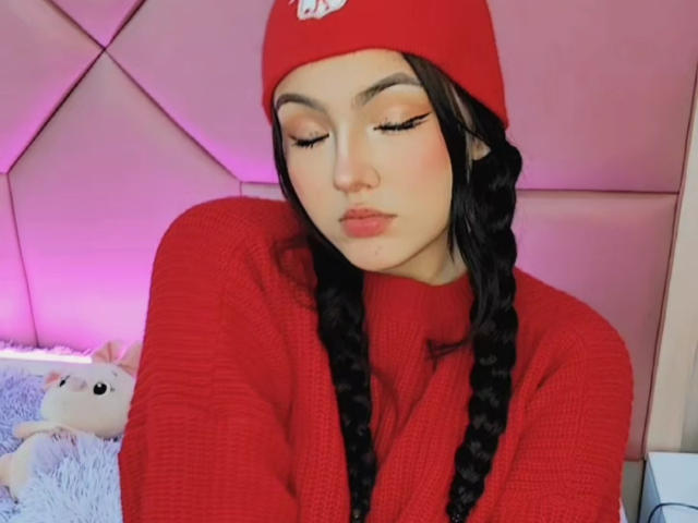 HaleiBeckham com longas tranças pretas, gorro vermelho e suéter vermelho, olhos fechados, iluminação rosada, parede rosa clara, um brinquedo de pelúcia branco ao fundo..