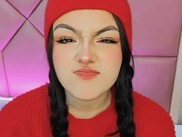 HaleiBeckham tem cabelos trançados pretos, usa um gorro vermelho e um suéter vermelho combinando, com sobrancelhas grossas, cílios longos e batom rosa, parecendo um pouco irritada..