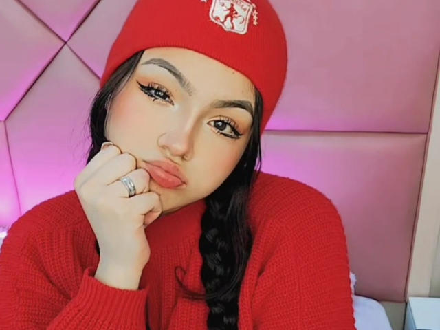 HaleiBeckham com lábios curvados, gorro vermelho e trança, vestindo um suéter vermelho, com a mão no queixo, um anel no dedo, um fundo iluminado em tons de rosa, delineador e um piercing no nariz..