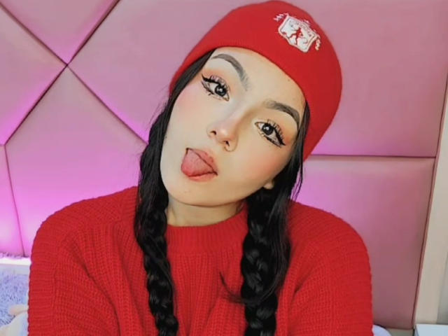 HaleiBeckham, com tranças longas e pretas, um gorro vermelho e um suéter vermelho, com a língua de fora, exibindo seu piercing no nariz e suas sobrancelhas grossas e sensuais..
