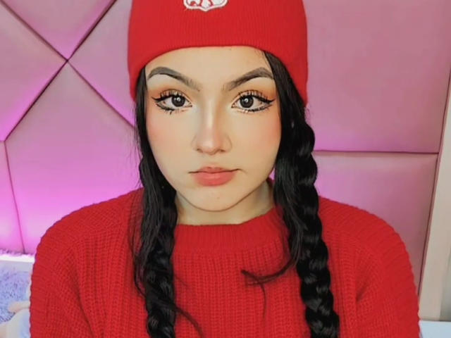 HaleiBeckham tiene la piel morena, cabello largo y negro trenzado, un gorro rojo de lana y un suéter rojo de punto. Está mirando directamente, con delineador de ojos alado y lápiz labial rosa. Sus senos no son visibles..