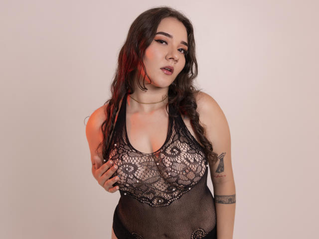 SofiSpinelli draagt een zwart kanten body, haar hand bedekt haar borsten, ze heeft golvend donker haar, een rode highlight, een tatoeage op haar rechterarm, en de achtergrond is wit. Ze kijkt licht geïrriteerd..