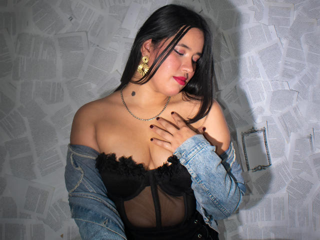 Rossy Granger mit schwarzen Haaren, rotem Lippenstift, trägt eine schwarze, durchsichtige Bluse, eine blaue Jeansjacke, goldene Stern-Ohrringe und eine Halskette, und berührt ihre Brüste mit Nagellack in Burgunderfarbe..