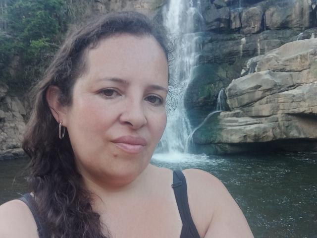 CandHoney ha i capelli castano ricci, la pelle chiara e piccoli orecchini. Indossa una canotta nera ed è in piedi vicino a una cascata, con un sorriso dolce sul viso..