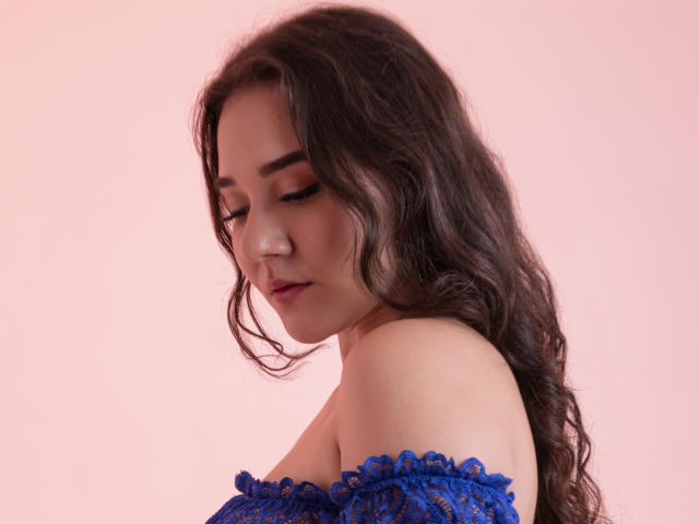 Sofi Spinelli, com cabelos castanhos ondulados, olhos fechados e uma blusa de renda azul com um ombro à mostra, revelando seus ombros e um sorriso discreto. Seus seios estão ligeiramente visíveis..