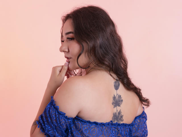 SofiSpinelli with wavy brown hair, blue lace off-shoulder top, finger to lips, showing a flower tattoo on her back, pink gradient background, soft expression.