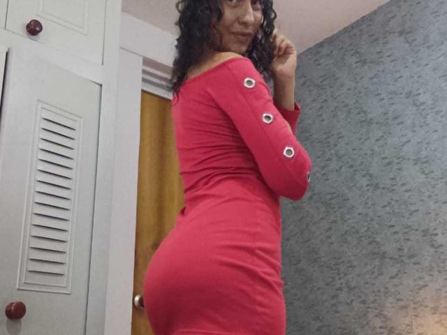 EroticSasha portant une robe rouge moulante avec des boutons, qui révèle ses fesses, et regardant par-dessus son épaule avec ses cheveux bouclés, souriant légèrement. Sa robe épouse ses courbes..