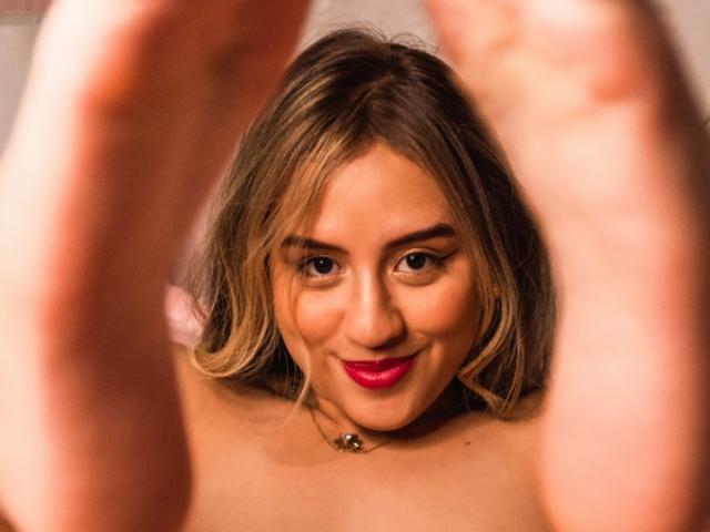 KylieSaoko ha i capelli biondi, il rossetto rosso e un sorriso carino, e guarda verso l'alto attraverso le sue mani alzate e sfocate, mostrando le spalle scoperte e una piccola collana..
