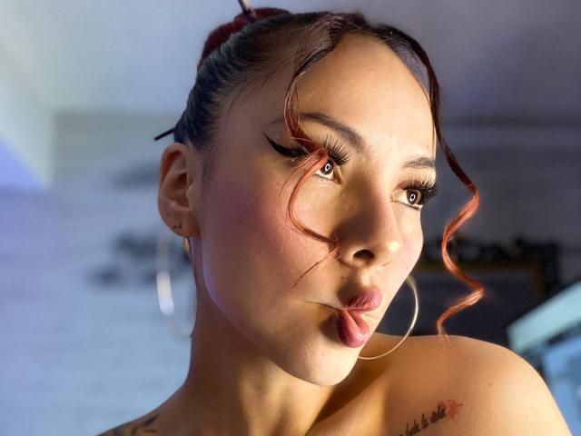 ValeryHarllow with pouty lips, brown eyes, and red curly hair, wearing gold hoop earrings, has a tattoo on her right shoulder and a cute, sexy face.