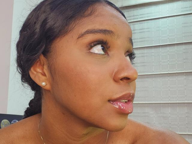 LisaMendez's face with shiny pink lips, dark brown hair in a braid, brown eyes looking up, wearing a diamond stud earring, and a thin gold necklace.
