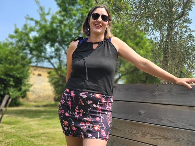 SexyAndFun, met een gemiddelde borstmaat, draagt een zwart topje zonder mouwen en een bloemenrok, staat bij een houten bank, glimlacht, draagt donkere zonnebril en een blauwe rugzak..