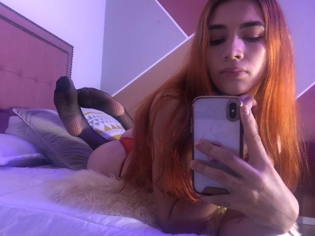Greta Harris, avec ses longs cheveux roux, est allongée sur un lit et prend un selfie avec son téléphone. Elle porte des bas résille et des sous-vêtements rouges, exposant ses fesses. La lumière de la chambre est violette..