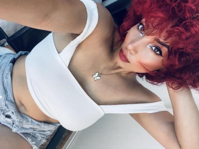 AmberDemons with curly red hair, white tank top, showing her tits, wearing denim shorts, and a butterfly necklace, looking up at the camera.