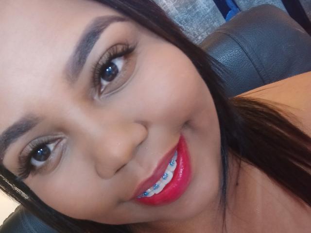 SarahSmity's face, showing brown eyes, dark eyebrows, long black hair, red glossy lips with blue braces, and a cute smile, against a black leather background.
