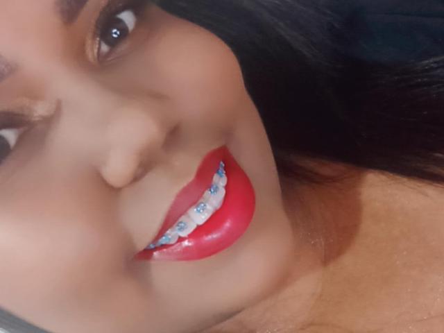SarahSmity's face with glossy red lips, white teeth with blue studs, and dark straight hair, smiling slightly. Her brown eyes are partially visible, looking to the left.