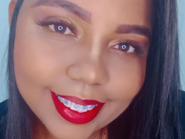 SarahSmity has tan skin, blue eyes, glossy red lipstick, and a slight smile showing her white teeth with blue braces. She has dark brown eyebrows and straight black hair.