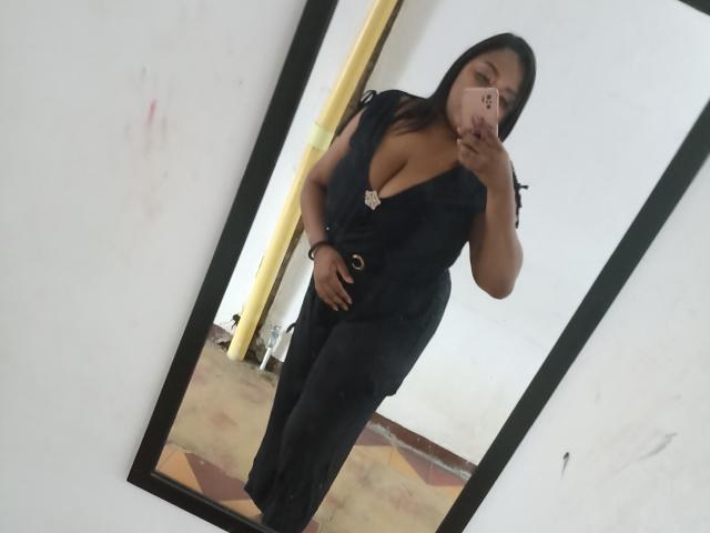 SarahSmity vestindo um vestido preto com um decote profundo, segurando o celular na frente de um espelho, mostrando seus seios e com a mão na virilha, em pé em um quarto..