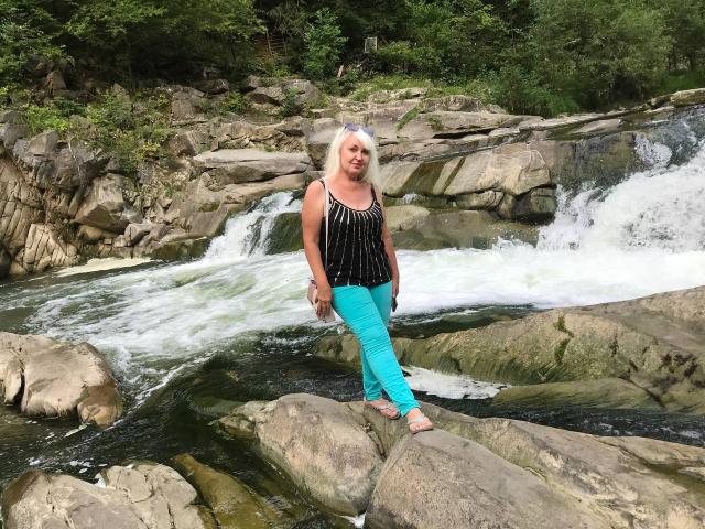 KamaliaSim mit weißem Haar, einem schwarz gestreiften Oberteil und hellblauen Hosen steht auf einem Felsen in einem fließenden Fluss und lächelt. Wasser stürzt um sie herum. Ihre Brüste sind klein..