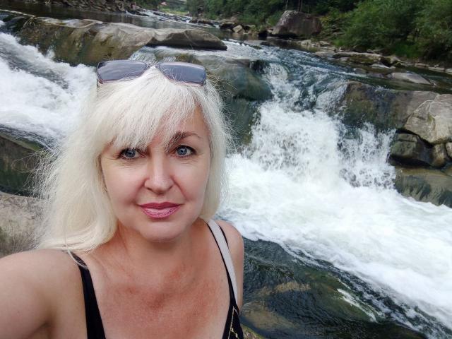 KamaliaSim has white hair, blue eyes, pink lipstick, black tank top, and sunglasses on her head, standing in front of a flowing waterfall. Boobs visible, smiling slightly.