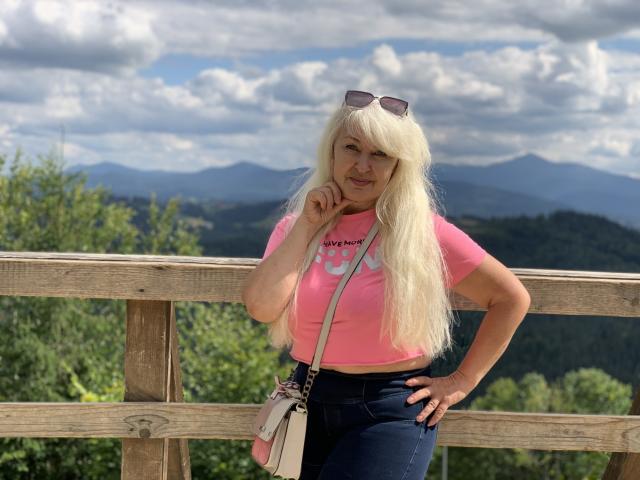 KamaliaSim has long blonde hair, pink t-shirt, and dark jeans, standing with a hand on her chin, wearing sunglasses on her head, and a white crossbody bag.
