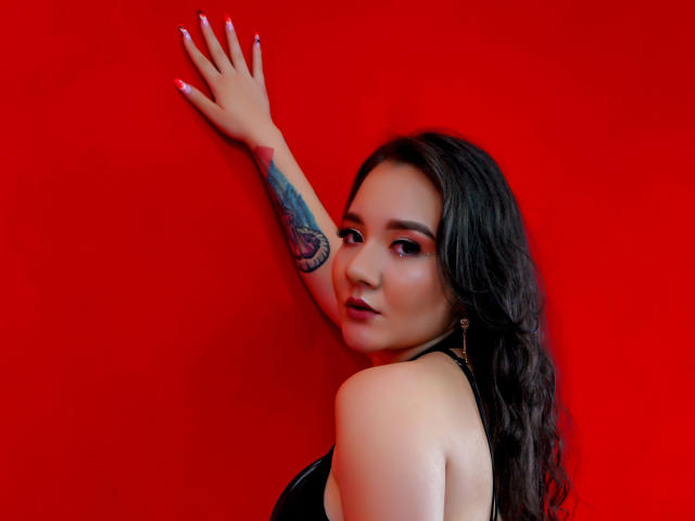 SofiSpinelli with long dark hair, looking back, arm raised, tattoo on arm, wearing black top, red background, white nails, slight smile, showing her tits, sexy.