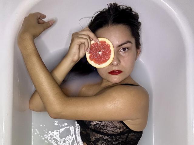 Jasmine Asha mit dunklem Haar, rotem Lippenstift und einem schwarzen Spitzenoberteil, die eine Grapefruit über ihrem rechten Auge hält, in einer weißen Badewanne, wobei Wasser spritzt..
