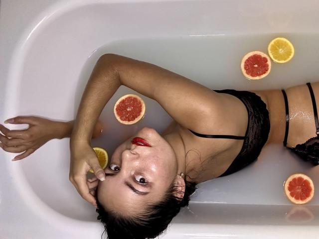 JasmineAsha is in a bathtub, wearing black lingerie, with her head back, looking up, red lipstick, wet black hair, and slices of grapefruit and lemon floating in the water.