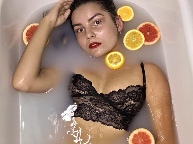 JasmineAsha in a white bathtub with water, wearing a black lace bralette, surrounded by sliced oranges, looking up with red lipstick, wet skin, and dark hair.
